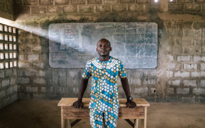 Young Togolese Secondary Education Teacher standing in front of a desk and chalk board, featuring a map of Africa